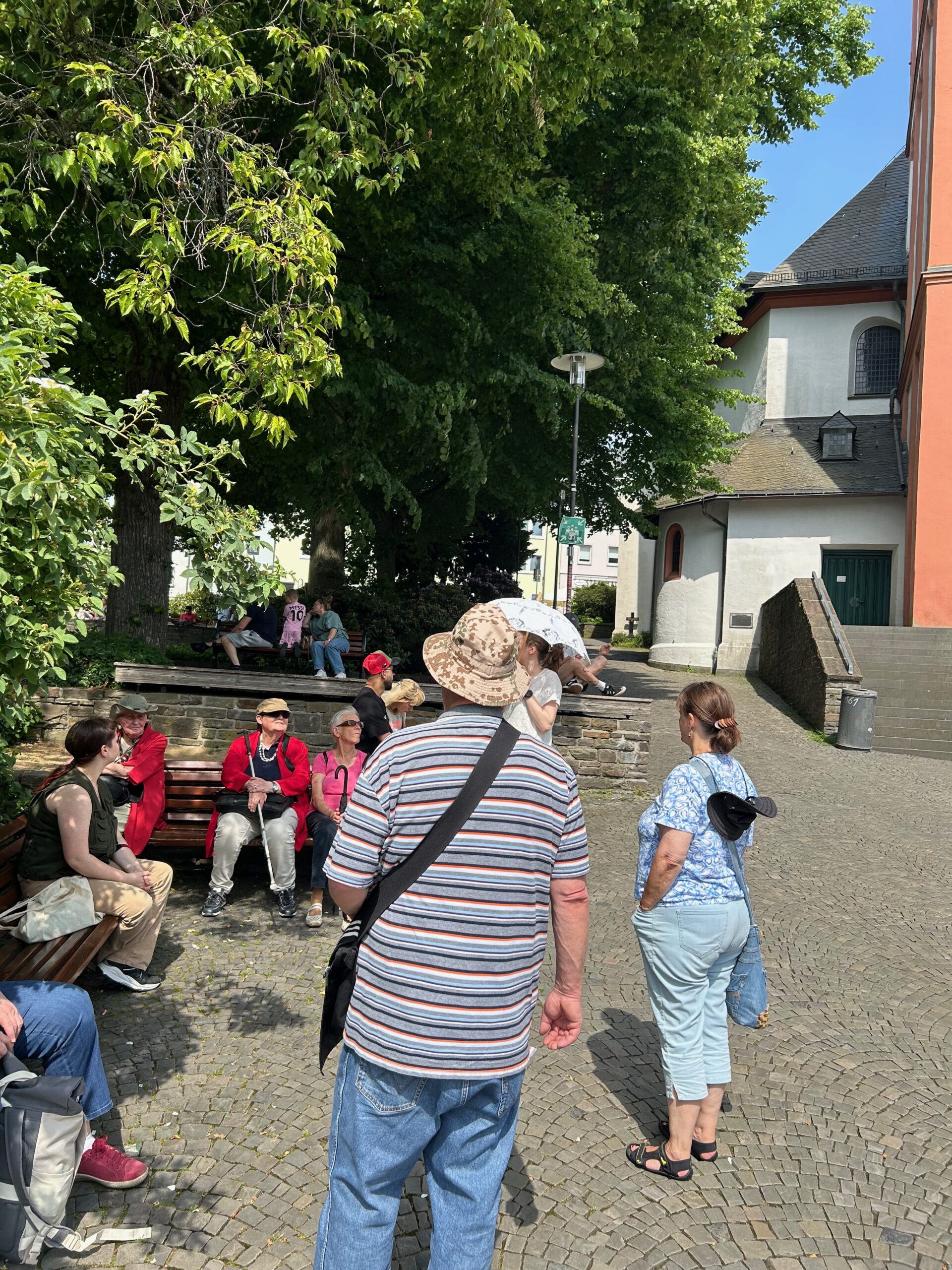 Ausflug Siegen, es sitzen Personen der besonderen Stadtführung im Schatten unter Bäumen vor der Martinikirche in Siegen. und hören der Stadtführerin zu.